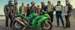 Groupe de motards souriants réunis autour d'une Kawasaki ZX6R verte brillante, partageant un moment convivial avec des accessoires humoristiques et un décor de route sinueuse en arrière-plan