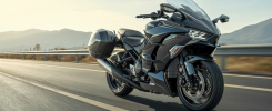 Moto sport-tourisme Kawasaki Ninja H2 SX SE 2025 noire avec superchargeur, tableau de bord TFT couleur 6,5 pouces, en pleine accélération sur route ouverte avec valises latérales et pare-brise réglable sous un ciel clair